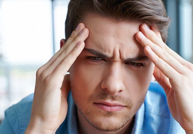Молодой мужчина в синей джинсовой рубашке и серой футболке сжимает виски обеими руками